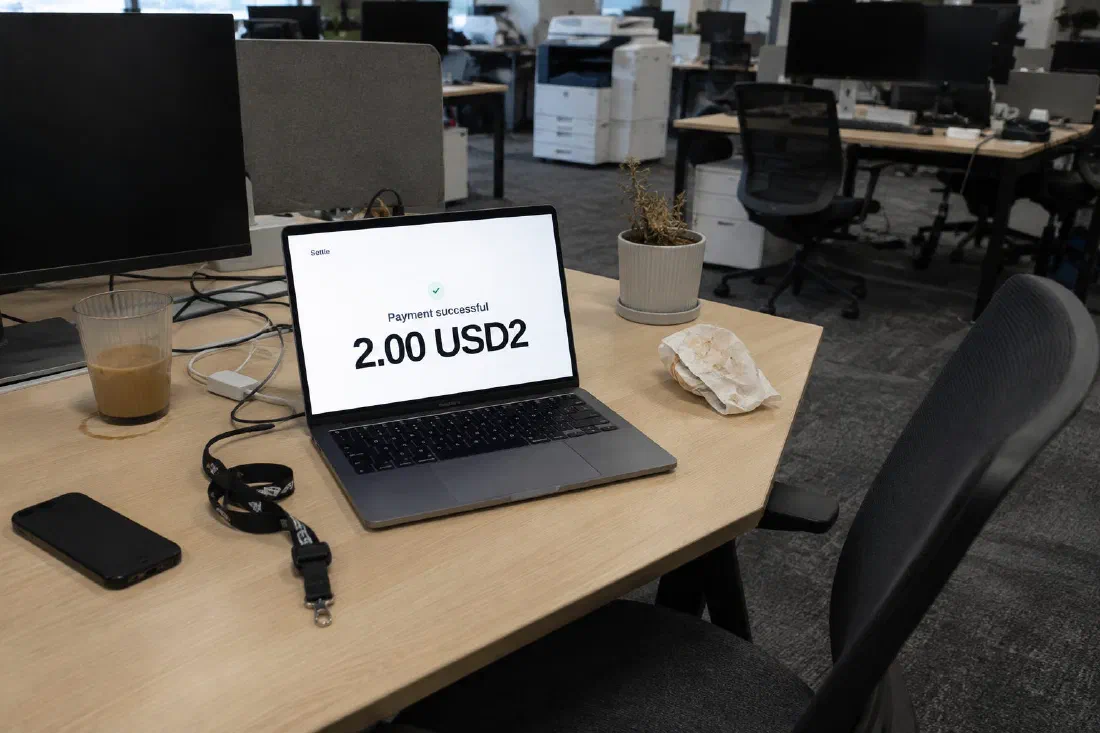 A laptop on an empty office desk beside a lanyard and a cold coffee. On screen: “Payment successful. 2.00 USD2.”