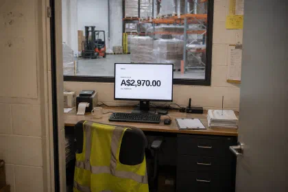 A warehouse office desk with a hi-vis vest over the chair. The monitor shows Pellinar’s “Agentic plan”, A$2,970.00, with the old price struck through beneath it.